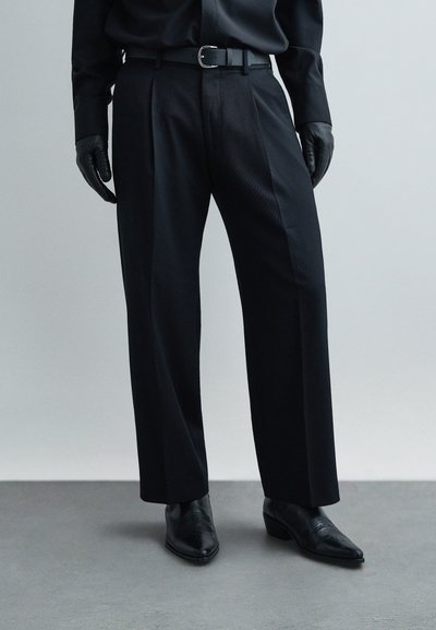 Pantalon noir sur mesure à coupe droite, tissu texturé et pli net, complété par une ceinture noire et des gants en cuir.