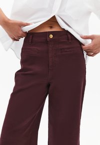 Pantaloni larghi bordeaux realizzati in tessuto morbido, con chiusura anteriore a bottone e due tasche posteriori, abbinati a una camicia bianca.