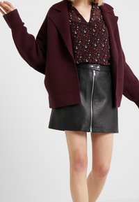 Burgundy blandad ull oversized jacka över en mönstrad svart och röd blus, kombinerad med en svart läder minikjol med dragkedja fram.