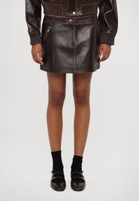Gonna mini in pelle marrone con tasca frontale con zip, design elegante e dettagli di cucitura visibili; abbinata a calze nere e scarpe bicolore.
