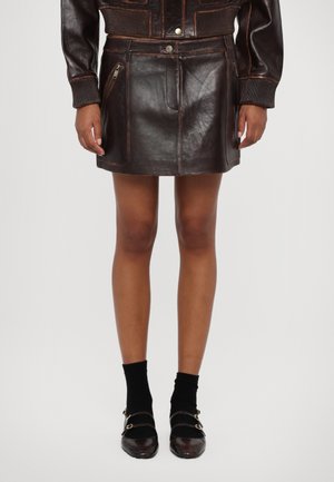 Gonna mini in pelle marrone con tasca frontale con zip, design elegante e dettagli di cucitura visibili; abbinata a calze nere e scarpe bicolore.
