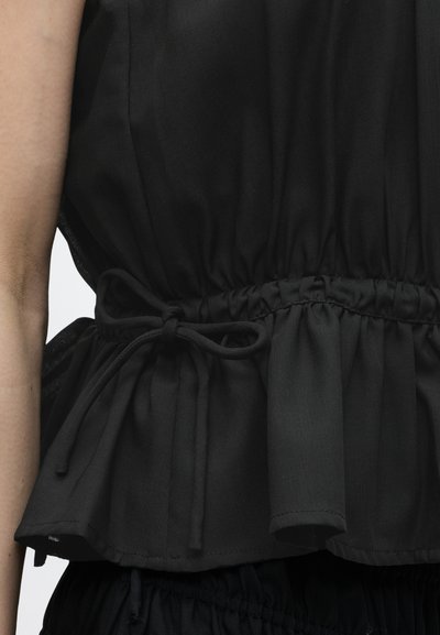 Κοντινό πλάνο σε μαύρο αμάνικο τοπ με δεμένη κορδόνι στη μέση και ραμμένο ύφασμα στο τελείωμα, φορεμένο από άτομο με ορατό χέρι.