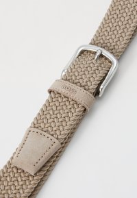 Vævet beige elastisk bælte med en sølvmetal spænde og en mærket ruskindsaftap med stikninger. Nærbillede af teksturen og designet.