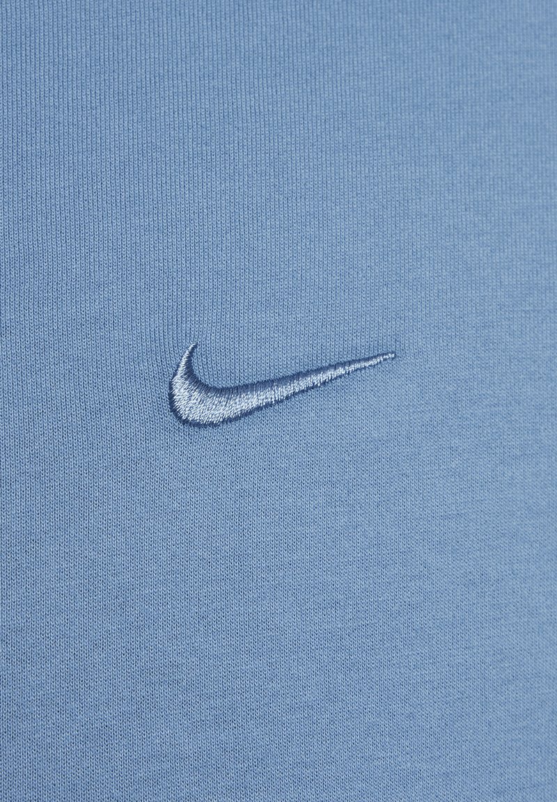 Jasnoniebieska bawełniana koszulka z małym, haftowanym białym logo Nike na lewej stronie. Gładka tekstura, prosty design.