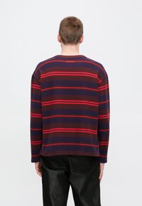 Μπλε σκούρο και μπορντό ριγέ μακρυμάνικο πουκάμισο με στρογγυλή λαιμόκοψη και κόκκινες λεπτομέρειες. Διαθέτει λογότυπο στην πλάτη κοντά στη λαιμόκοψη.