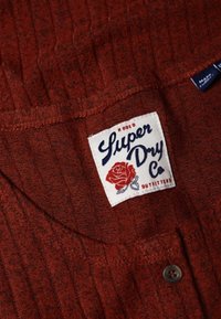 Tissu rouge à nervures avec une étiquette blanche indiquant "Super Dry Co" et un graphique de rose rouge, accompagné d'un bouton sombre sur le vêtement.