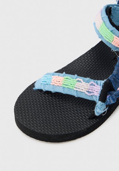 Niebieska materiałowa sandała z teksturowaną czarną podeszwą. Posiada kolorowy, pleciony pasek z pastelowymi koralikami. Projekt obejmuje postrzępione krawędzie i detale szwów.