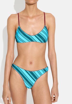 Kobieta ubrana w niebiesko-turkusowy bikini w paski z cienkimi bordowymi ramiączkami, stojąca na tle jednolitego jasnego tła.