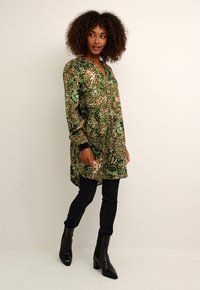 Blouse à manches longues verte et beige avec des motifs floraux et animaliers, associée à un jean skinny noir et des bottines noires à talons pointus.