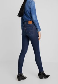 Jeans ajustados de mezclilla azul oscuro, con un parche de cuero marrón en la parte trasera y costuras clásicas. Combinados con una camisa de mezclilla y botas negras.