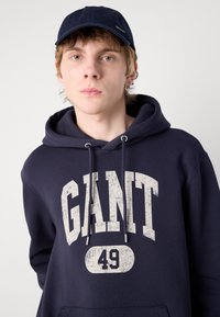 Marineblå hettegenser med stor slitt hvit "GANT 49"-trykk og snor. Brukt med en matchende marineblå caps. Glatt stofftekstur.