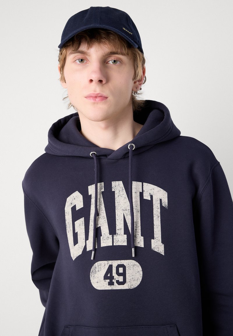 Felpa blu navy con una grande stampa bianca "GANT 49" invecchiata e cordino. Indossata con un cappellino blu coordinato. Texture del tessuto liscia.