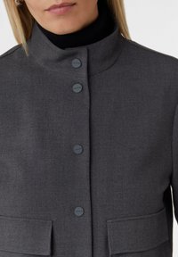 Kobieta w ciemnoszarej, dopasowanej marynarce z wysokim kołnierzem, czarnymi guzikami z etykietą "comma" i kieszeniami klapowymi, nosi czarny golf.