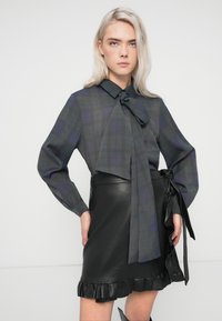 Ruudullinen paita tummassa vihreässä ja laivastonsinisessä värissä, jossa on suuri solmio kauluksessa, yhdistettynä mustaan nahkaiseen minihameeseen, jossa on röyhelöreuna.