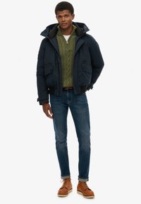 Granatowa kurtka puchowa z odpinanym kapturem, kieszenie boczne; zielony sweter z warkoczowym wzorem; niebieskie dżinsy; brązowe sznurowane buty ze szwami w kontrastowym kolorze.