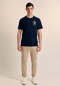 Camiseta de algodón azul marino de manga corta, con detalles de logotipo y cuello redondo, combinada con pantalones beige remangados y zapatillas blancas.