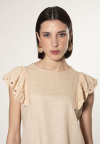 Top in lino beige con delicate maniche a volant caratterizzate da dettagli in pizzo. Una rifinitura in pizzo accentua i bordi delle maniche.