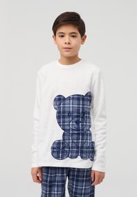 Biała polarowa koszula z długim rękawem z niebieskim wzorem niedźwiedzia w kratę. Granatowe piżama w kratę dopełniają zestaw, pokazując miękki, teksturowany materiał.