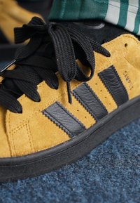 Yellow suede sneaker with black leather stripes and laces. Features a textured upper and a black rubber sole. Close-up view of the shoe.