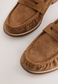 Mocassins en daim marron avec une couture détaillée et une semelle plate, présentés en gros plan, vue de dessus, sur un fond neutre.