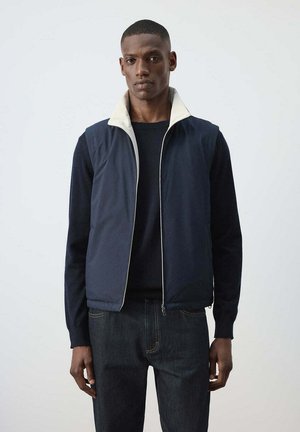 Hombre que lleva un chaleco navy azul con cremallera y cuello blanco sobre una camiseta negra de manga larga y vaqueros oscuros, de pie frente a un fondo liso y claro.