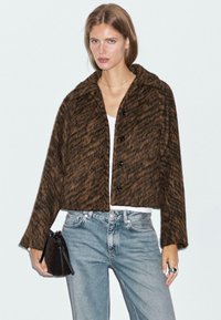 Veste courte à motifs marron et noir avec un col, boutons noirs et manches longues, portée sur un haut blanc et associée à un jean bleu.