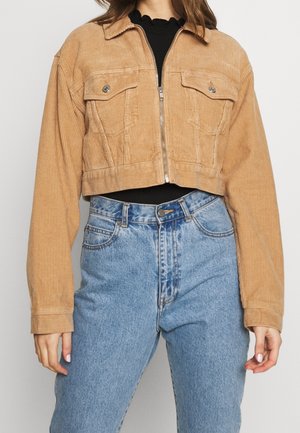 Femme portant une veste courte en velours côtelé beige avec poches avant, par-dessus un top noir, associée à un jean bleu taille haute.