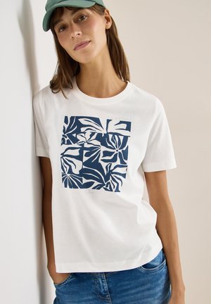Femme portant un t-shirt blanc avec un motif de feuilles abstraites bleu marine et un jean bleu, appuyée contre un mur clair, portant une casquette verte.