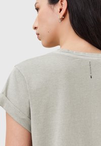 T-shirt grigia chiara con maniche arrotolate, texture morbida e dettaglio del logo vicino al colletto sul retro. Design semplice e minimalista.