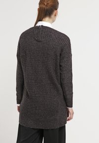 Tummanharmaa neulottu villapaita, jossa on resorikuviointi, pitkät hihat ja vetoketjusuljenta takana. Kaareva helma lisää ainutlaatuista muotoa ja designia.