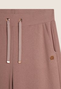 Pantaloni della tuta rosa chiaro realizzati in tessuto morbido, dotati di un cinturino con cordino e dettagli oro, oltre a una tasca laterale.