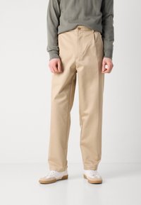 Beige bomullbyxor med rak skärning, med dragkedja och knappstängning, tillsammans med vita sneakers och en grå långärmad topp.