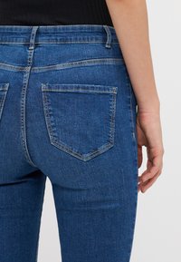 Blue denim jeans featuring a fitted design, rear pockets with distinct stitching, and a smooth texture with a slight stretch.