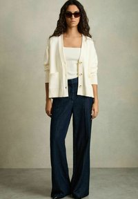 Cardigan crema con polsini a coste e due tasche frontali, abbinato a jeans a gamba larga blu scuro. Capelli lunghi e mossi e occhiali da sole scuri completano il look.