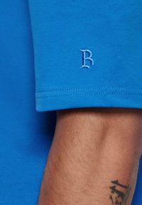 Blå bomulds T-shirt med rund halsudskæring, korte ærmer og et lille broderet "B" logo nær ærmeenden. Glat tekstur.