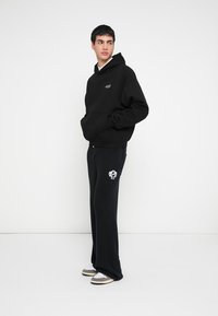 Fekete kapucnis pulóver kenguru zsebbel, mellrészen logóval, bordázott fekete szabadidőnadrággal, amely grafikával díszített. Hozzáillő laza lábbeli.