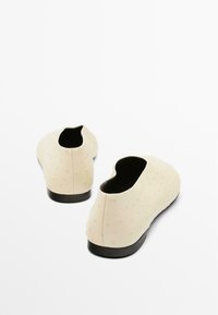 Scarpe slip-on beige con superficie testurizzata e piccole protuberanze, tacco basso e design aperto sul retro. Suola nera visibile sul fondo.