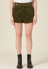 Olivgrüne Cordshorts mit hoher Taille, Taschen vorne und zwei dekorativen Knöpfen. Kombiniert mit schwarzen Knöchelschuhen.