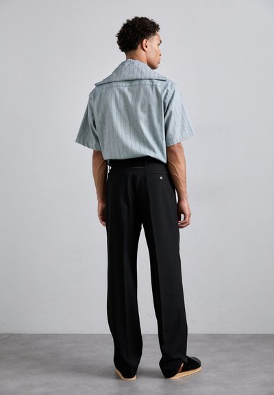 Camicia a maniche corte di colore azzurro chiaro con righe verticali, abbinata a pantaloni neri su misura e scarpe con plateau in legno.