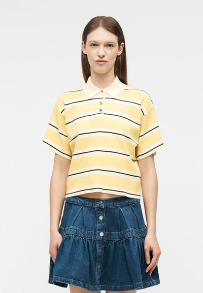 Γυναίκα στέκεται όρθια, στραμμένη προς τα εμπρός, φορώντας μπλούζα πόλο με κοντά μανίκια κίτρινη και λευκή με ρίγες και μια μπλε τζιν φούστα με κουμπιά.
