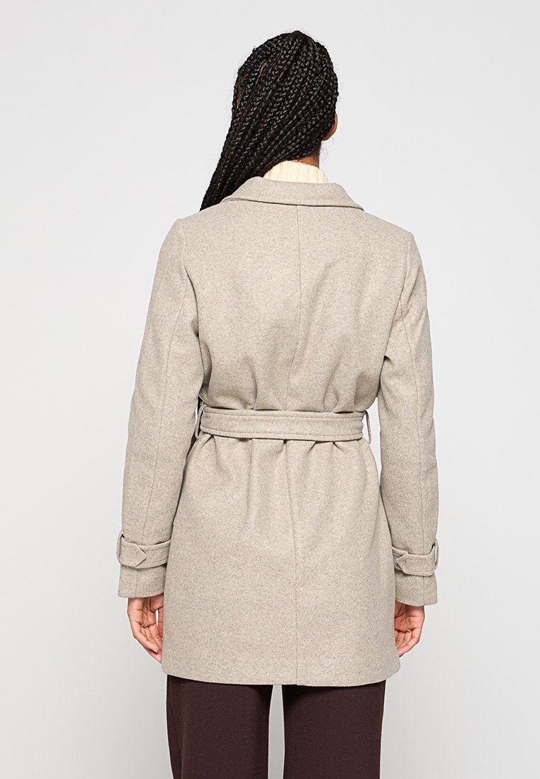 Femme avec de longs cheveux tressés portant un manteau beige ceinturé, se tenant de dos contre un fond clair.