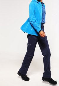 Blauwe softshell jas met een capuchon en ritszakken, gecombineerd met donkere marineblauwe broeken. Zwarte wandelschoenen met gestructureerde zolen maken de outfit compleet.