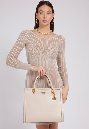 Borsa tote in pelle beige con hardware dorato, forma rettangolare, etichetta decorativa e design strutturato, tenuta da una persona in un vestito aderente.