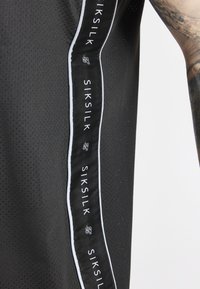 Svart atletisk linne med nätstruktur, med vitt SIKSILK-märke på en sidopanel. Kantat med kontrasterande vit piping.