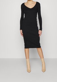 Robe noire à manches longues avec décolleté en V, coupe ajustée et longueur jusqu'aux genoux. Portée avec des bottines pointues couleur nude. Texture lisse.