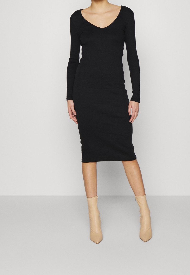 Robe noire à manches longues avec décolleté en V, coupe ajustée et longueur jusqu'aux genoux. Portée avec des bottines pointues couleur nude. Texture lisse.