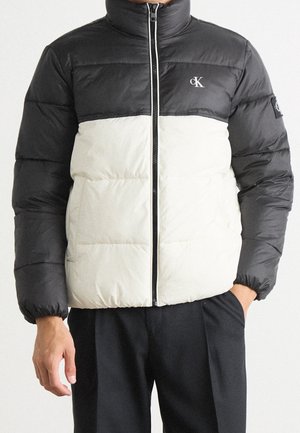 Homme portant une veste matelassée noire et blanche avec le logo CK sur la poitrine et la manche, debout avec une main dans la poche.