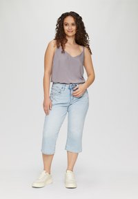 Haut sans manches gris clair avec un col en V, associé à un jean cropped bleu clair. Le modèle se tient en baskets blanches devant un fond uni.