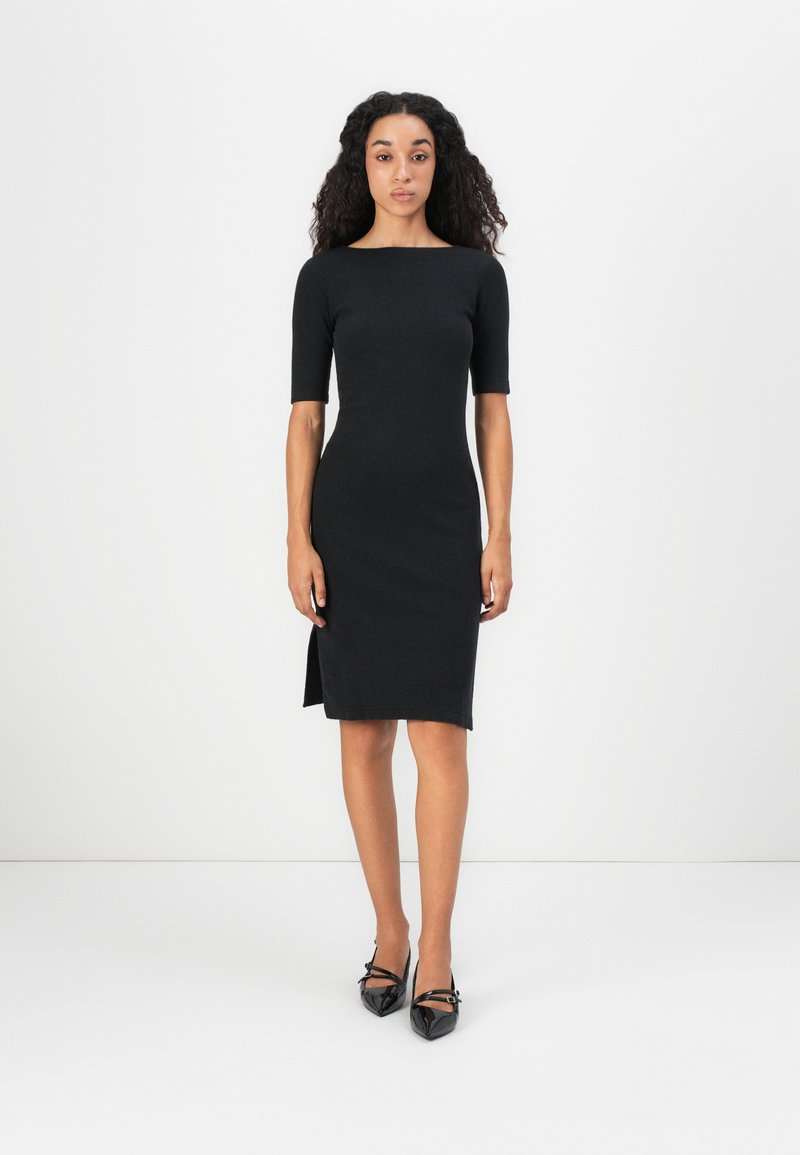 Black fitted dress with short sleeves and a boat neckline. Features a side slit and textured fabric, paired with black patent heels.