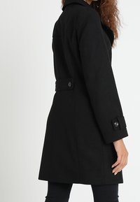 Svart ullkappa med en strukturerad silhuett, slagkrage och knappdetaljer på ärmar och midja. Sllät textur, knälång design.